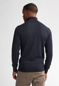 Pullover blu scuro con un colletto alto, polsini e orlo a coste, realizzato in tessuto a maglia liscia. Indossato con pantaloni beige.