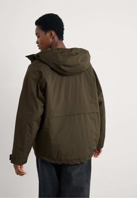 Oliwkowa kurtka z kapturem o luźnym kroju, wykonana z lekkiego materiału. Posiada obniżony tylny brzeg i regulowane mankiety.