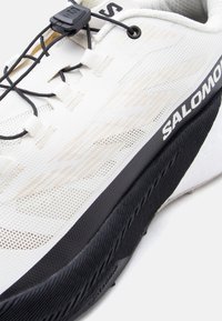 Λευκό αθλητικό παπούτσι Salomon με μαύρη σόλα, μαύρο σύστημα γρήγορου δέσιμου και υφαντό επάνω μέρος με υφή, με μπεζ σχέδια και λογότυπο.