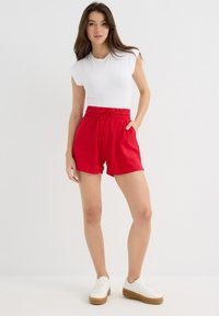 Pantalones cortos rojos elásticos con una cinturilla ancha y cordón. Incluye dos bolsillos laterales. Combinado con una camiseta blanca ajustada y zapatillas blancas.
