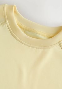 Gros plan d’un t-shirt jaune pâle à col rond montrant une double couture au niveau du col et un détail de bande tressée cousue au niveau du cou.