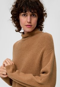 Brauner, gerippter Strickpullover mit Rollkragen, lockerem Schnitt, überschnittene Schultern und langen Ärmeln; weiche Textur. Das Model posiert mit gefalteten Händen.