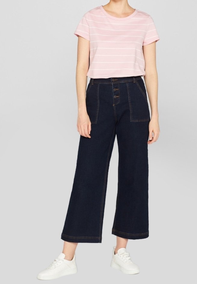 Femme portant un t-shirt rose à manches courtes rayé, un jean foncé taille haute à jambes larges avec des boutons sur le devant, et des baskets blanches, debout sur un fond gris.