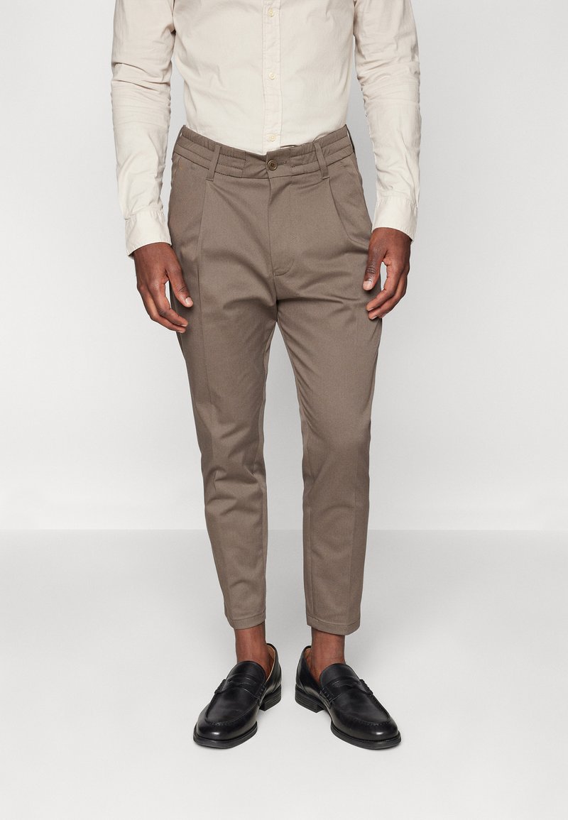 Pantalones marrones ajustados con una textura sutil, que cuentan con un cierre frontal de botón y trabillas para cinturón, combinados con mocasines negros.