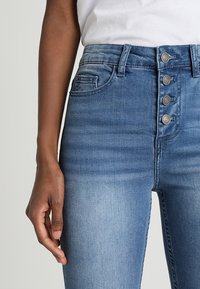 Vyšší modré džíny z denimu s vybledlým vzhledem, vybavené pěti knoflíky na zip, poutky na pásek a předními kapsami. Hladká textura s mírným elastickým efektem.