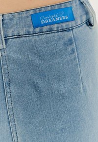 Svetlomodré denimové džínsy s hladkou textúrou a výraznou modrou etiketou s nápisom "Sunlight DREAMERS" blízko pásu.