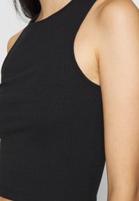 Zbliżenie osoby w czarnej, prążkowanej, bezrękawnikowej bluzce crop top, ukazującej ramię i górną część klatki piersiowej.