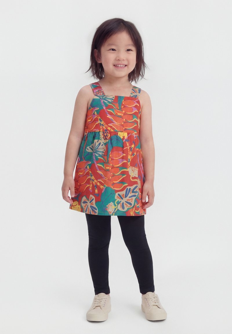 Χαμογελαστή κοπέλα που φοράει πολύχρωμο φλοράλ αμάνικο φόρεμα, μαύρα κολάν και μπεζ αθλητικά παπούτσια, στέκεται μπροστά σε μια απλή λευκή φόντο.