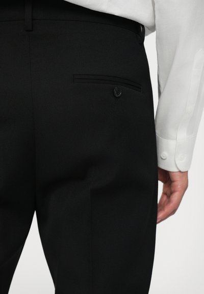 Pantaloni negri tailorați cu o textură netedă, având un buzunar posterior cu un nasture asortat. O cămașă albă este parțial vizibilă deasupra.