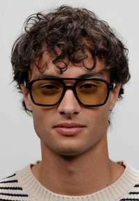 Lunettes de soleil écaille de tortue marron avec des verres teintés jaunes, design à monture carrée, arborant des accents argentés sur les branches. Le modèle porte un pull rayé.