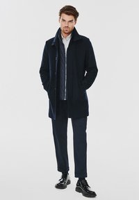 Granatowy płaszcz wełniany z domieszką, z klapkowym kołnierzem, bocznymi kieszeniami i zapinaną na zamek wewnętrzną warstwą. Połączony z ciemnymi spodniami i błyszczącymi czarnymi butami.