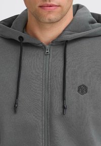 Hoodie gris zippé en matériau doux. Doté de cordons de serrage noirs, d'un petit logo brodé et d'une fermeture éclair lisse avec un accent orange.