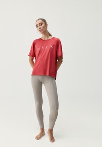 Červené sportovní tričko s krátkým rukávem a světle šedými textovými designy, kombinované s těsnými béžovými legínami, hladká textura a bez viditelných vzorů.