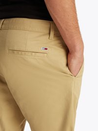 Chinos bege feitos de uma mistura de algodão, apresentando um corte plano, bolsos laterais e um pequeno patch de marca com detalhes em vermelho, branco e azul.