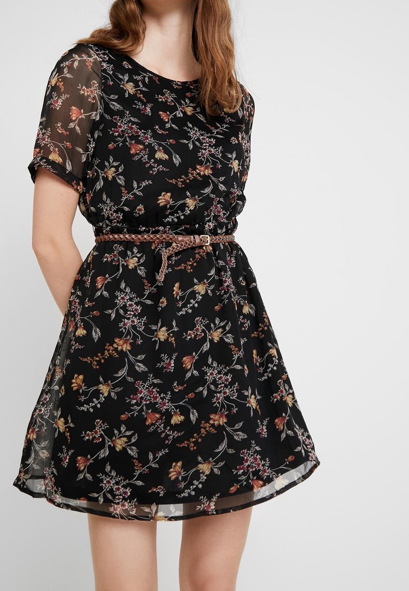 Femme portant une robe courte noire à fleurs avec des manches transparentes et une ceinture tressée marron à la taille sur un fond uni.