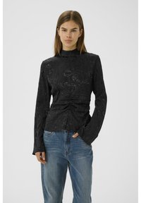 Haut noir à manches longues avec un col haut, motif texturé en tourbillon et détails froncés à la taille, associé à un jean bleu clair.