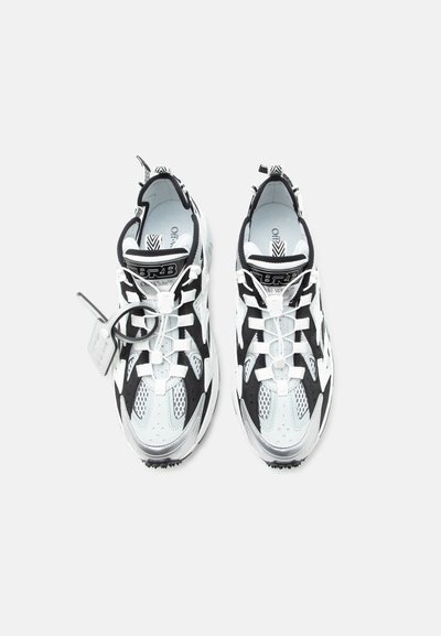 Ζευγάρι αθλητικών παπουτσιών σε μαύρο, λευκό και ασημί χρώμα με λεπτομέρειες από δίχτυ, λογότυπα στη γλώσσα και προσαρτημένη πλαστική ετικέτα, απεικονιζόμενα από πάνω σε λευκό φόντο.