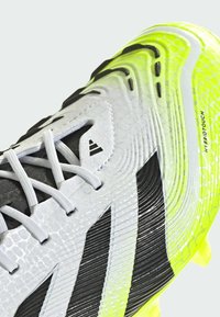 Soccer cleat in white and black with a textured surface and neon yellow accents. Features a lace-up closure and pronounced outsole studs.