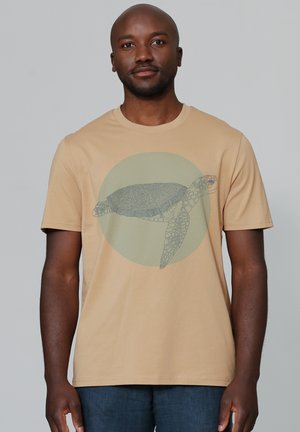Beige T-shirt met korte mouwen, voorzien van een grafische afbeelding van een schildpad op een groene cirkelachtige achtergrond. Gemaakt van een zachte katoenmix en heeft een ontspannen pasvorm.