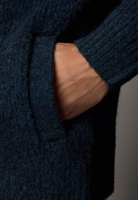 Pulover tricotat de culoare bleumarin, cu un finisaj texturat. Prezintă o buzunar lateral, manșete tricotate și o croială lejeră. Prim-plan cu o mână în buzunar.