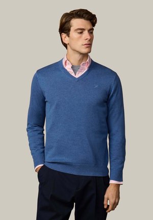 Man draagt een blauwe V-hals trui over een roze overhemd met een gestreept onderhemd, staat met één hand in de zak tegen een effen achtergrond.