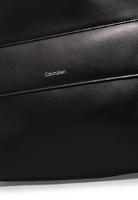Borsa in pelle nera con cuciture sottili, texture liscia e logo bianco "Calvin Klein". Design minimalista con una silhouette elegante.