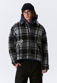 Jongere in een zwarte beanie, zwart-grijs geruite zip-up jack, marineblauwe hoodie, zwarte broek, bril en een kettingaccessoire.