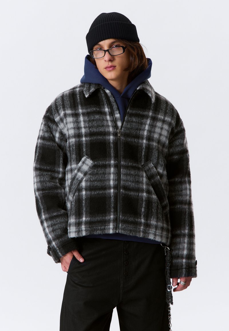 Jongere in een zwarte beanie, zwart-grijs geruite zip-up jack, marineblauwe hoodie, zwarte broek, bril en een kettingaccessoire.