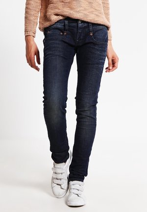 Person trägt dunkelblaue Slim-Fit-Jeans, beigen Strickpullover und weiße Sneaker und geht vor einem schlichten weißen Hintergrund nach vorne.