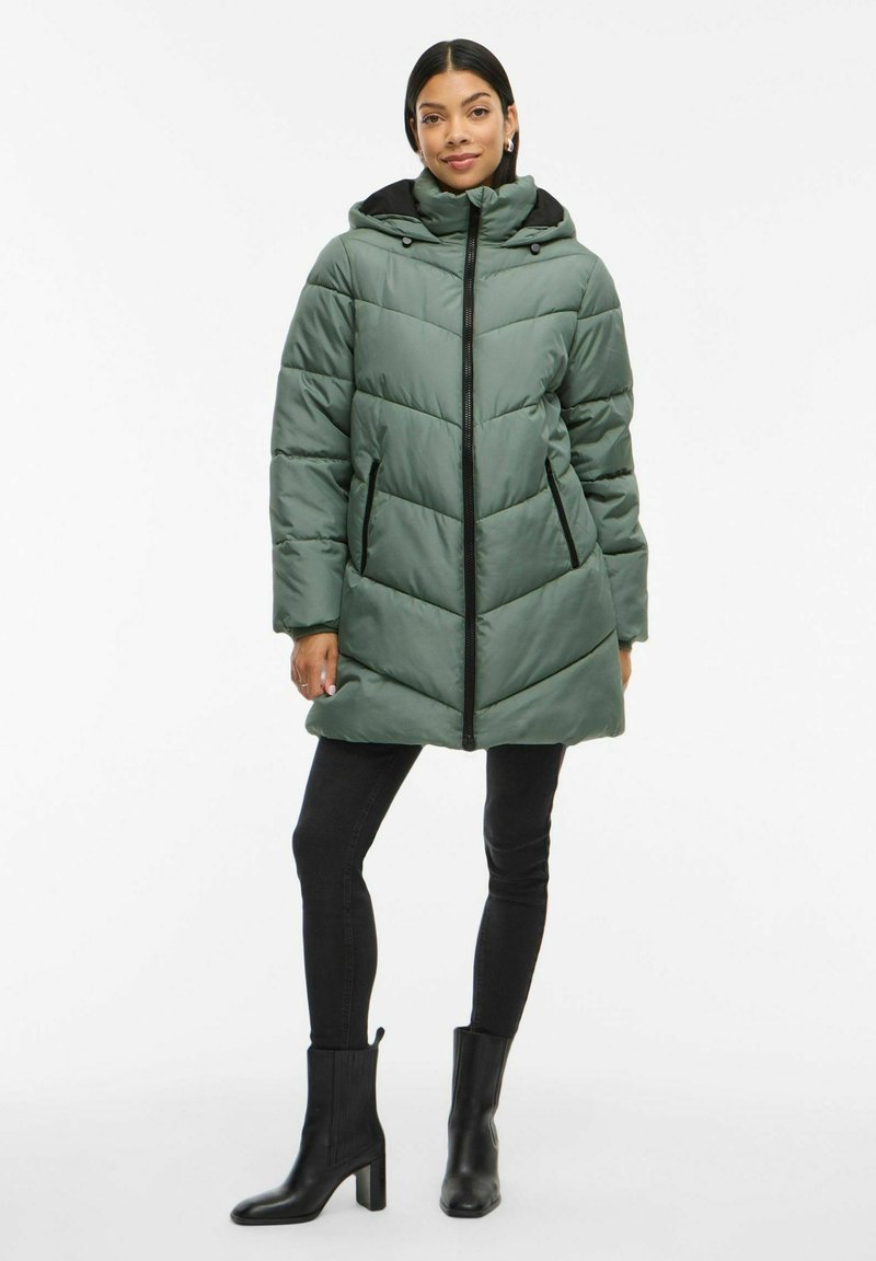 Polstret grøn parka med lynlås foran, chevron-syning, justerbar hætte og sorte detaljer. Båret over sorte skinny bukser og ankelstøvler.