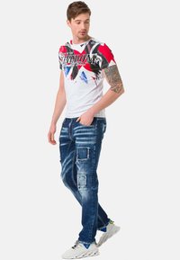 Herren-Grafik-T-Shirt in Weiß mit roten, blauen und schwarzen abstrakten Mustern, kombiniert mit einer verwaschenen dunkelblauen Jeans mit Patch-Akzenten.