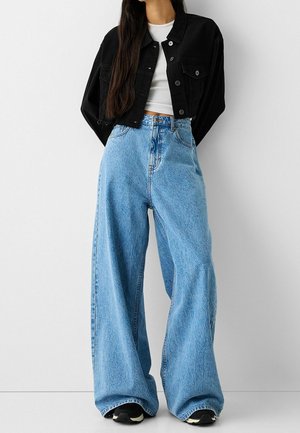 Vrouw met lichtblauwe hoog getailleerde jeans met wijd uitlopende pijpen, zwart cropped jasje, witte crop top en zwarte sneakers tegen een effen witte achtergrond.