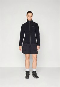 Ung person som står, iført svart glidelås-fleecejakke, svarte cargo-shorts, hvite sokker og svarte tursko mot en ensfarget bakgrunn.