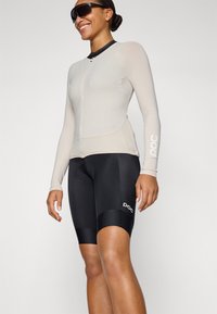 Lys beige langermet sykkeltrøye med glidelås foran, kombinert med svarte polstrede sykkelshorts med hvit logo.
