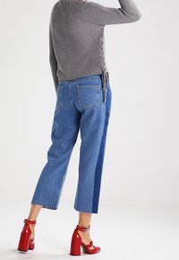 Grijze geribbelde trui met veterdetails aan de zijkanten, gecombineerd met blauwe denim cropped pants met een contrasterend zijpaneel en een rafelrand. Rode hoge hakken sandalen.