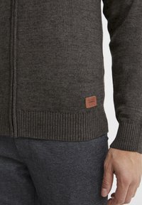 Suéter de punto gris oscuro con puños y dobladillo acanalados, que presenta una pequeña etiqueta de marca de cuero marrón en la parte inferior izquierda. Textura suave.
