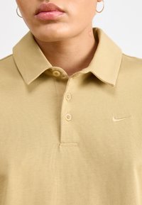 Polo shirt in cotone color sabbia con colletto e tre bottoni. Presenta un piccolo logo ricamato sul lato sinistro del petto. Texture liscia, design casual.