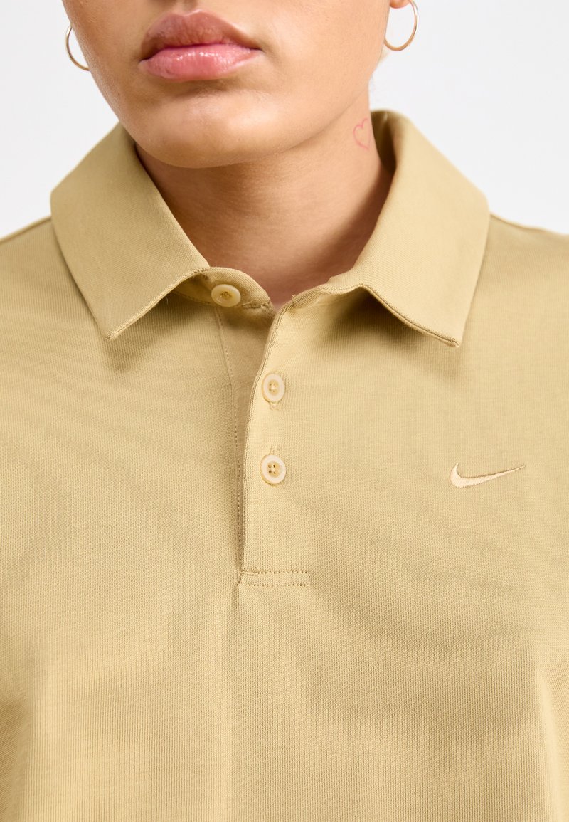 Polo en coton beige avec un col et trois boutons. Présente un petit logo brodé sur la poitrine gauche. Texture lisse, design décontracté.