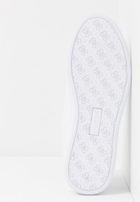 Semelle de sneaker blanche avec un motif texturé affichant un design de logo répétitif et une étiquette rectangulaire indiquant "GUESS". Le bord est lisse.
