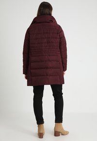 Giacca imbottita borgogna con collo alto, design trapuntato verticale e taglio diritto, abbinata a pantaloni neri e stivaletti beige.