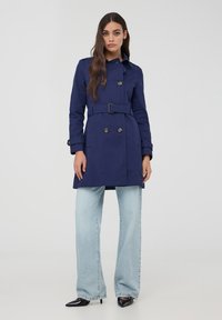 Cappotto trench blu navy con bottoni doppi, vita con cintura e tasche laterali, abbinato a jeans a gamba larga blu chiaro e tacchi neri.