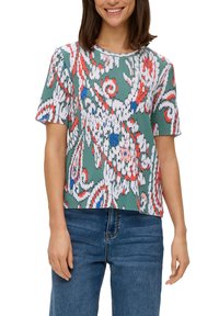 Kobieta w krótkim rękawku z kolorowym abstrakcyjnym wzorem w zielonym, białym, czerwonym, niebieskim i różowym, nosząca niebieskie dżinsy.