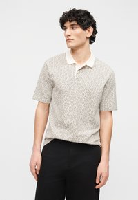 Νέος άνδρας με σγουρά σκούρα μαλλιά, φορώντας κοντομάνικο πόλο με σχέδια και μαύρο παντελόνι, κοιτάζοντας προς τα αριστερά του πάνω σε ένα απλό φόντο.