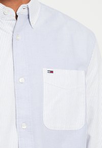 Ανοιχτό μπλε πουκάμισο με κουμπιά, με ριγωτό γιακά και τσέπη αριστερά στο στήθος. Μαλακή υφή, αντίθετο ριγέ σχέδιο στην τσέπη.