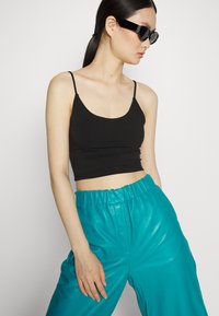 Top cropped preto com alças finas, combinado com calças de imitação de pele brilhante em teal de cintura alta. Modelo a usar óculos de sol escuros.