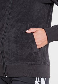 Giacca zippata nera realizzata in tessuto morbido e testurizzato. Presenta polsini a coste e una tasca frontale, sottolineando il comfort e la funzionalità.