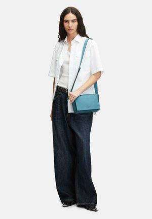 Taschen aus tealfarbenem Leder in einer strukturierten Form, mit einer strukturierten Oberfläche und verstellbarem Riemen. Das Modell trägt ein weißes Hemd und dunkle Jeans.