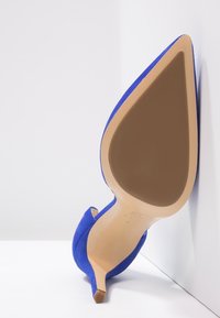 Modrá sametová stiletto bota, špičatá špička, úzký podpatek, béžová podrážka a hladký povrch. Minimalistický design bez viditelných ozdob.