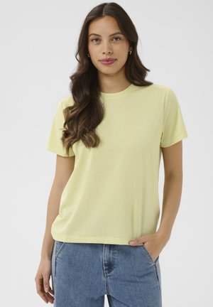 Jeune femme aux longs cheveux ondulés portant un t-shirt jaune pâle à manches courtes et un jean bleu, debout avec une main dans la poche.