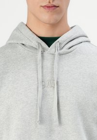 Un sweat à capuche gris avec un cordon de serrage, présentant une texture douce, un logo brodé au centre, superposé sur une chemise verte.